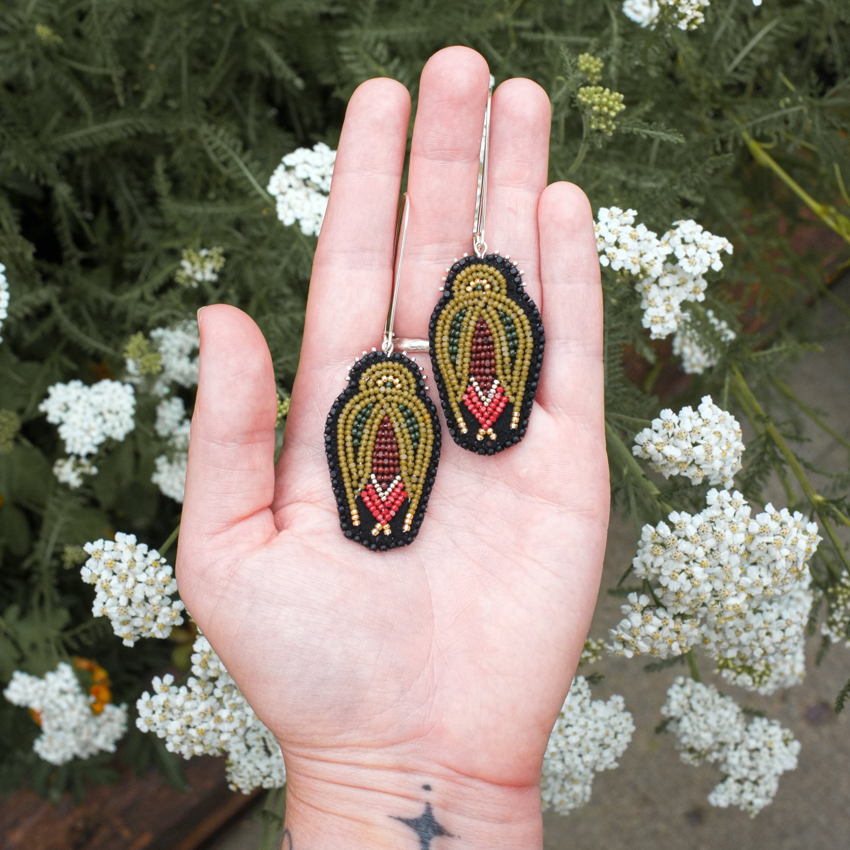 A person's hand holding a pair of handmade beaded earrings with a black, gold, and red design, set against a background of green foliage and small white flowers.